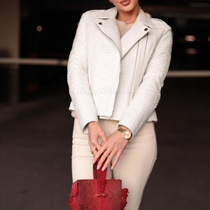 Puede incluir: Una chaqueta de cuero blanco texturizado con un dise&ntilde;o cl&aacute;sico de cremallera asim&eacute;trica. La chaqueta se combina con pantalones color crema y un bolso de mano rojo. Se ve un reloj dorado en la mu&ntilde;eca. El estilo general es elegante y moderno.
