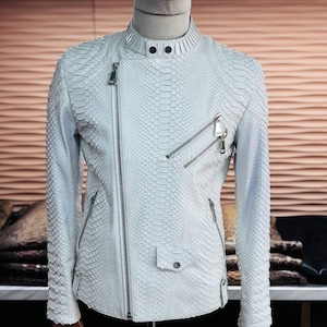 Pode incluir: Casaco de couro branco, com textura de pele de cobra, com gola alta e múltiplos zíperes prateados. O casaco tem uma silhueta ajustada e um pequeno bolso com fecho de pressão. O casaco é exibido em um manequim.