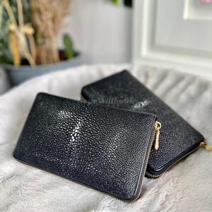 Puede incluir: Dos carteras negras con una superficie texturizada y moteada y cremalleras doradas. Las carteras son rectangulares y parecen ser de cuero. Están sobre una superficie suave y clara.