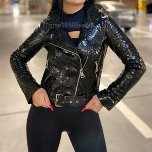 Op de afbeelding: Zwarte leren jas met slangenpatroon, ritssluiting en riem met gouden gesp. De jas heeft een klassieke bikerstijl met een reverskraag en lange mouwen. De tekst "HopeWillbe" is zichtbaar op de jas.