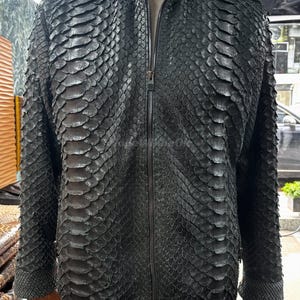 Può includere: Giacca in pelle nera con motivo a scaglie e chiusura a zip. La giacca presenta un collo alto e maniche lunghe. L'indumento è esposto su un manichino.