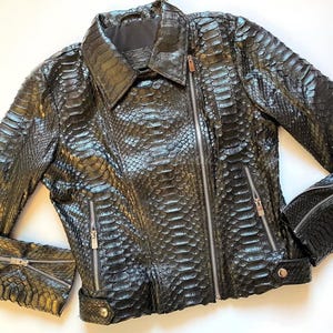 Pode incluir: Casaco de couro preto com padrão de pele de cobra. O casaco tem gola, fecho de correr e vários bolsos com fecho. As mangas têm fechos nos punhos. O casaco é de cor escura com um ligeiro brilho.