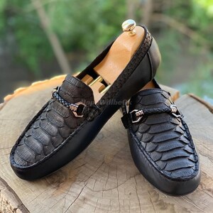 May include: A pair of black leather loafers with a snakeskin pattern. The shoes have a braided strap across the top with a gold-colored buckle. Wooden shoe trees are inside the shoes.