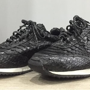 May include: A pair of black sneakers with a textured, scaly pattern, possibly snakeskin. The shoes have white soles and black laces. The sneakers are designed for casual wear.