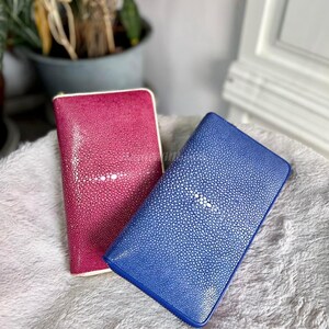 May include: Two rectangular wallets. One is a deep pink with a white border, and the other is blue. Both wallets have a textured surface with a central white design. The wallets are displayed on a soft, white surface.
