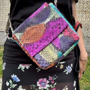 Pode incluir: Uma bolsa tiracolo de formato quadrado com um padrão de pele de cobra texturizada em tons vibrantes de magenta, laranja, roxo, amarelo e azul-petróleo. A bolsa possui fecho de aba e alça a condizer, com um design geométrico multicolorido.