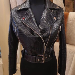 Może przedstawiać: Czarna skórzana kurtka z wzorem skóry węża. Kurtka ma kołnierz z klapami, asymetryczne zapięcie na zamek i pasek w talii. Ma długie rękawy i dopasowaną sylwetkę. Widoczny jest tekst "Hope4meOK".