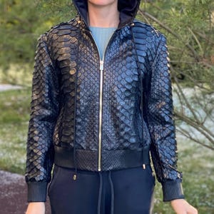 Può includere: Giacca con cappuccio nera con motivo a scaglie di rettile. La giacca presenta una cerniera dorata, coulisse nera e polsini e orlo a coste. La giacca è indossata con pantaloni neri.