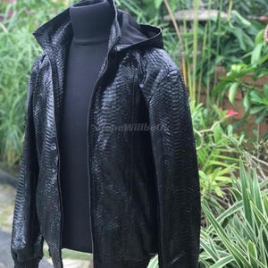 May include: Black hooded jacket with a textured snakeskin pattern. The jacket has a full zip, a high collar, and ribbed cuffs and hem. The jacket is displayed on a black turtleneck.