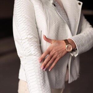 Puede incluir: Chaqueta de cuero blanca texturizada con un patr&oacute;n de piel de serpiente. La chaqueta tiene un cuello cl&aacute;sico, cierre de cremallera y mangas largas. Se ve un reloj dorado en la mu&ntilde;eca. Prenda de vestir exterior elegante.