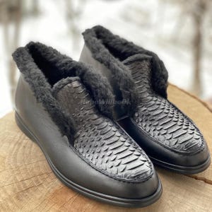 May include: A pair of black leather shoes with a snakeskin pattern on the vamp. The shoes have a soft, black fur lining around the opening. The shoes are sitting on a wooden surface.