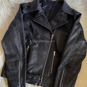 May include: Black leather jacket with a snakeskin texture. The jacket features a notched collar, an asymmetrical zipper closure, and multiple zippered pockets. A belt with a silver buckle is at the waist. The jacket is displayed on a white fur-like surface.