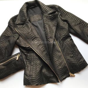 May include: A dark gray, textured leather jacket with a snake-skin pattern. The jacket features a notched collar, an asymmetrical zipper closure, and a zippered pocket. The jacket has long sleeves and a tailored fit.