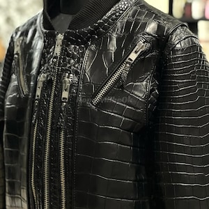 May include: A close-up of a black leather bomber jacket with a crocodile skin texture. The jacket has multiple silver zippers and a ribbed collar. The jacket is displayed on a mannequin.