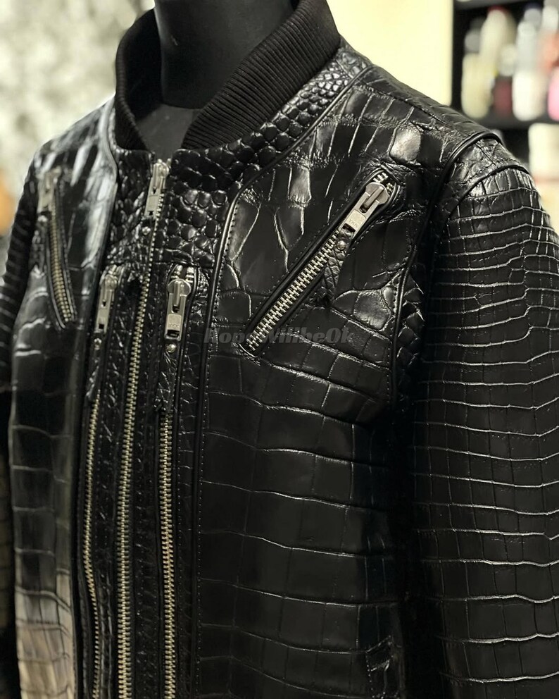 May include: A close-up of a black leather bomber jacket with a crocodile skin texture. The jacket has multiple silver zippers and a ribbed collar. The jacket is displayed on a mannequin.
