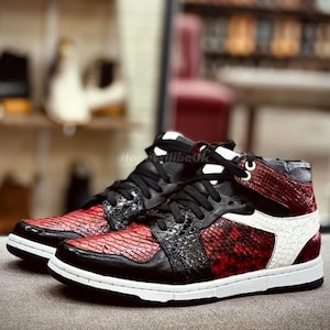 May include: High-top trainers in black, red, and white. The red sections have a scale-like texture, while the black areas are smooth. The trainers have white soles and black laces. The design is eye-catching and modern.