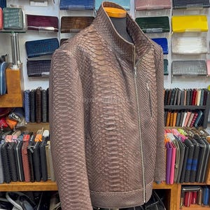 May include: A brown snakeskin-patterned leather jacket with a zip closure and a stand-up collar. The jacket is displayed on a mannequin. The background shows shelves filled with wallets and bags in various colours.