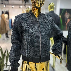 Può includere: Giacca bomber grigio scuro con un motivo strutturato simile alla pelle di serpente. La giacca presenta una cerniera argentata, polsini e orlo a coste neri. Esposta su un manichino dorato.