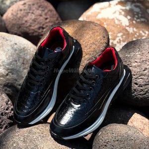 May include: A pair of black sneakers with a textured, reptile-like pattern. The shoes have a white sole and red interior lining. The sneakers are laced up and sit on a bed of rocks.