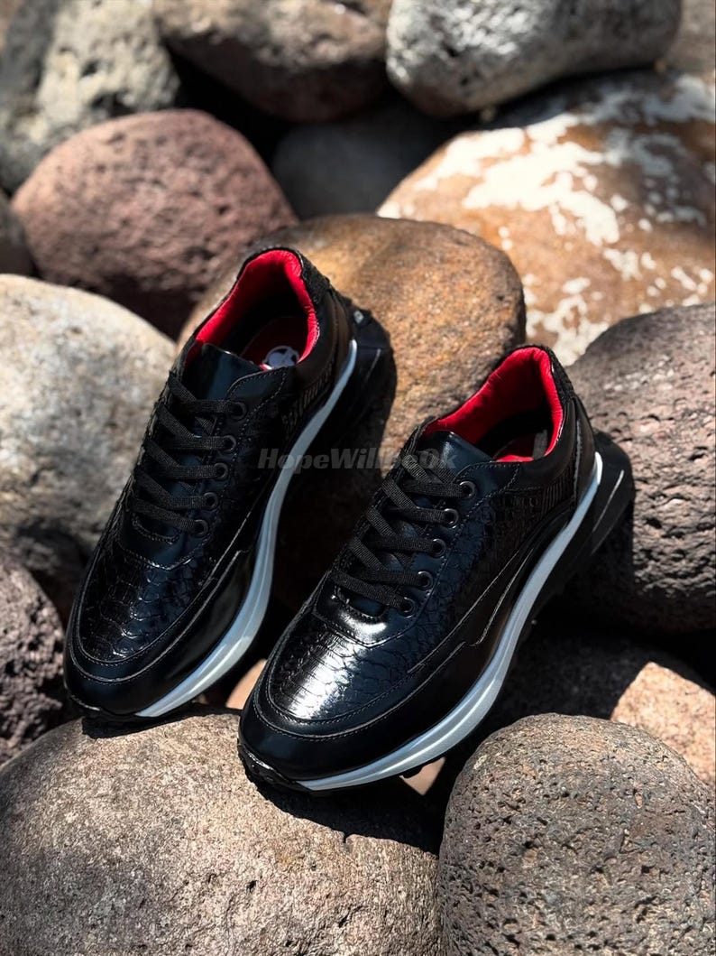 May include: A pair of black sneakers with a textured, reptile-like pattern. The shoes have a white sole and red interior lining. The sneakers are laced up and sit on a bed of rocks.