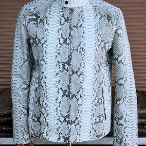 May include: A light-coloured jacket with a snakeskin pattern. The jacket has a stand-up collar, a zip closure, and two side pockets. The snakeskin pattern features shades of white, grey, and black. The jacket is displayed on a mannequin.