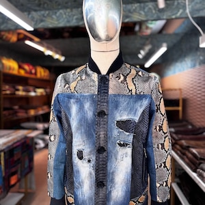 Può includere: Giacca bomber in denim e pelle di serpente su un manichino. La giacca presenta un davanti in denim blu con dettagli strappati e bottoni neri. Le maniche e la parte superiore della schiena sono decorate con un motivo a pelle di serpente marrone, beige e nero.