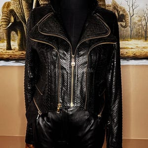 Può includere: Giacca in pelle nera con texture a pelle di serpente e cerniere dorate. La giacca ha un colletto alto e multiple tasche con cerniera. La giacca è esposta su un manichino. Lo sfondo presenta un dipinto di elefanti.