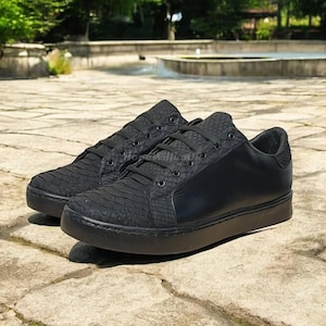 May include: A pair of black trainers with textured panels and smooth leather accents. The shoes have black laces and a thick, solid black sole. The trainers are displayed on a stone surface.