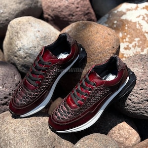 Könnte beinhalten: Ein Paar weinrote Sneaker mit schwarzen Schnürsenkeln und einem strukturierten Muster an Zehen und Seiten. Die Sneaker haben eine weiße Sohle und eine schwarze Ferse, vor dem Hintergrund großer, abgerundeter Steine.