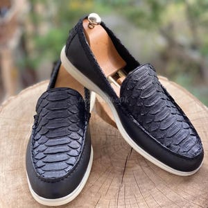 Puede incluir: Un par de mocasines de cuero negro con textura de piel de serpiente en la pala. Los zapatos tienen suela blanca y se exhiben sobre una superficie de madera. Zapatos de estilo sin cordones.