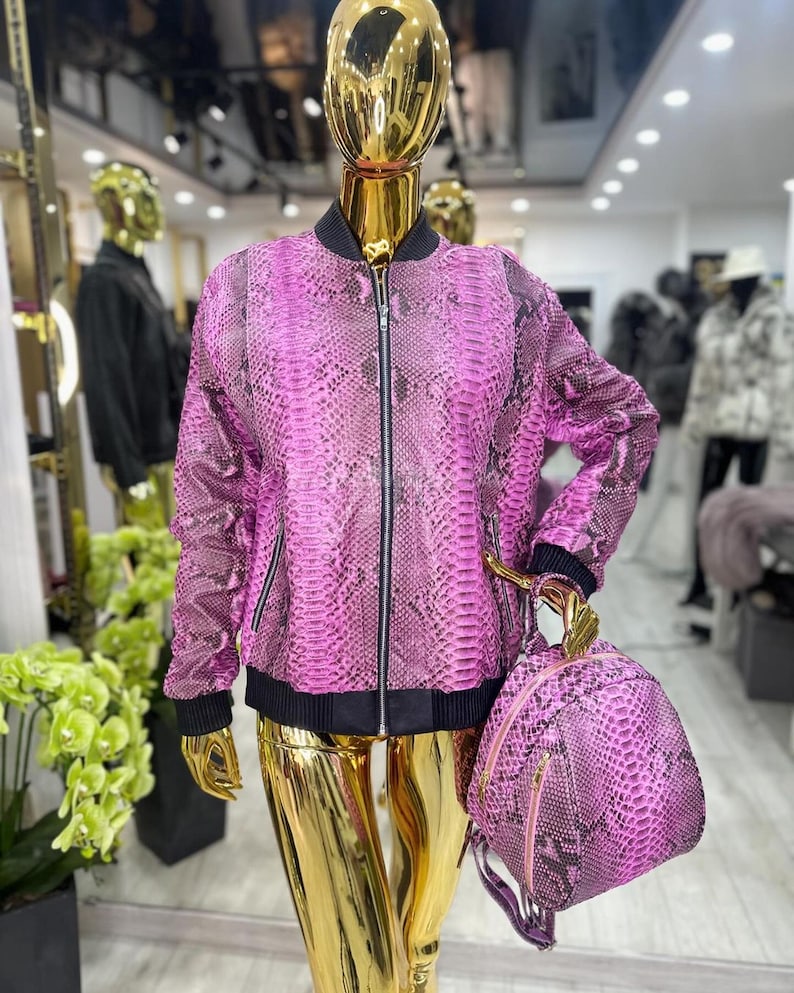 May include: A pink snakeskin-effect bomber jacket with a black trim and zip, presented on a gold mannequin. A matching backpack with gold detailing is also shown. The jacket and backpack are the main focus of the image.