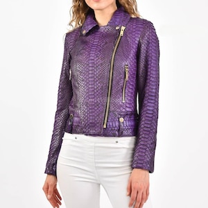 May include: A purple leather jacket with a snakeskin pattern. The jacket has a notched collar, an asymmetrical zipper, and two zippered pockets. The jacket has gold-colored zippers and hardware. The model is wearing white pants.