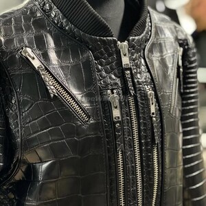 May include: Black leather bomber jacket with a crocodile skin texture. The jacket has multiple silver zips and a ribbed collar. The sleeves have a striped design. The jacket is displayed on a mannequin.