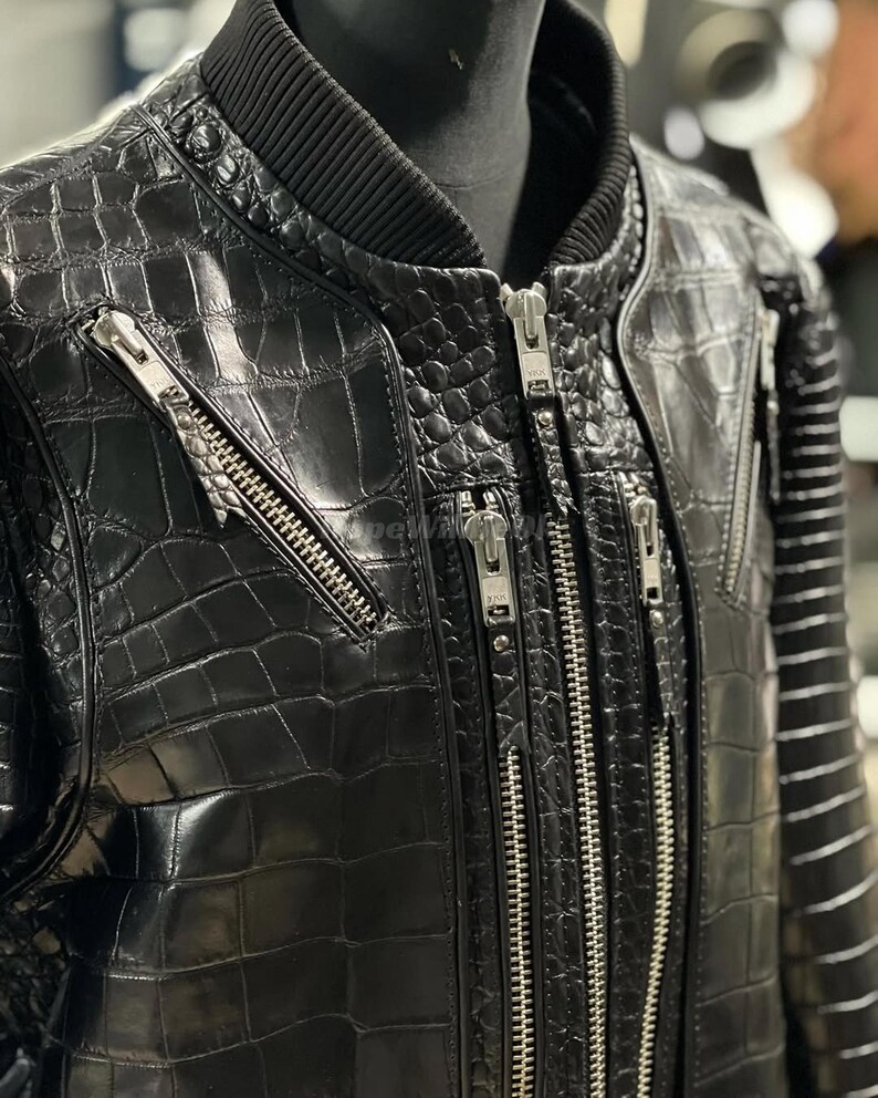 May include: Black leather bomber jacket with a crocodile skin texture. The jacket has multiple silver zips and a ribbed collar. The sleeves have a striped design. The jacket is displayed on a mannequin.
