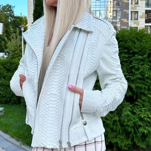 Pode incluir: Casaco de couro branco texturizado, com padrão de pele de cobra. O casaco de estilo motociclista tem gola entalhada, vários zíperes e bolsos. A modelo usa o casaco com calças xadrez.