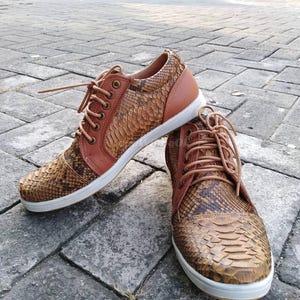 Könnte beinhalten: Ein Paar braune und beige Sneaker mit Schlangenmuster. Die Schuhe haben braune Schnürsenkel, beige Lederakzente und weiße Sohlen. Die Sneaker sind lässig und stilvoll.