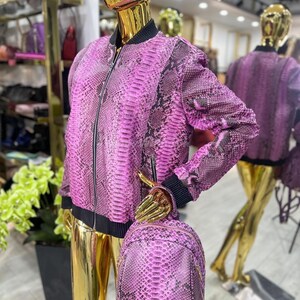 May include: A pink and black snakeskin-patterned bomber jacket and matching backpack. The jacket features a black collar and cuffs, and a zip closure. The backpack has adjustable straps. The items are displayed on a gold mannequin.