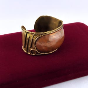 May include: A wide cuff bracelet featuring a polished copper centre panel framed by textured brass edges. The brass border includes linear detailing and a small circular accent, creating a mixed-metal design with a warm, metallic finish and open back.