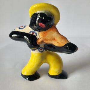 May include: A ceramic figurine of a musician in a yellow outfit and hat, black shoes and arms, and a cream-colored shirt with blue and red circles. The musician's face is black with red lips.