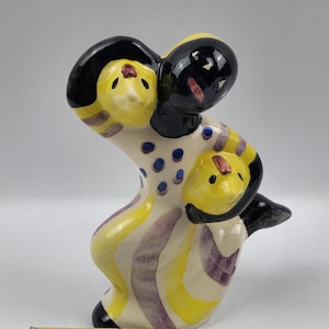 May include: A ceramic figurine depicting three stylized figures. The figures have yellow faces with black hair and are embracing. The figurine is painted with yellow, purple, and blue accents on a cream background. The overall design is whimsical and artistic.