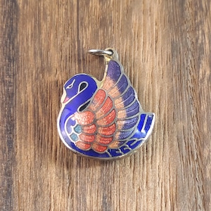 May include: A silver-toned swan pendant with blue, orange, and purple enamel detailing. The swan has a curved neck and outstretched wings, with a small loop at the top for a chain. The pendant is set against a wooden background.