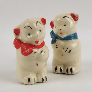 May include: Two vintage ceramic bear figurines. The bears are off-white with red accents on their ears and mouths. One bear wears a red scarf, and the other wears a blue scarf. The bears have black painted details and blue eyes.
