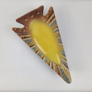 May include: A vintage ashtray in a unique arrowhead shape. The ashtray features a central yellow basin, surrounded by a brown border with textured details. The edges are decorated with blue and yellow patterns, creating a retro aesthetic.
