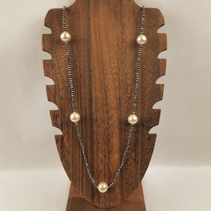 May include: A long necklace featuring large, cream-colored faux pearls interspersed with small, dark gray beads. The necklace is displayed on a wooden jewelry stand, showcasing its elegant design and potential for accessorizing various outfits.
