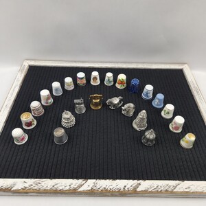 May include: A collection of decorative thimbles arranged on a black felt board within a white-framed tray. The thimbles feature various designs, including floral patterns, buildings, and animals. Some are silver-toned, while others have colorful painted details.