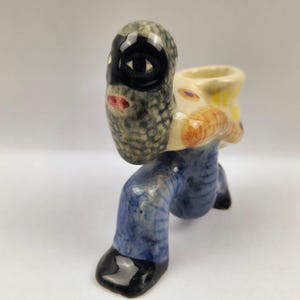 May include: A ceramic figurine depicting a person in a black mask and blue pants, with a beige and yellow sack on their back. The figurine has a unique, artistic design with a glossy finish.