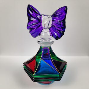 May include: A decorative glass perfume bottle with a purple bow-shaped stopper. The bottle has a geometric design with red, blue, and green panels, accented with gold detailing. The base is a light blue colour.
