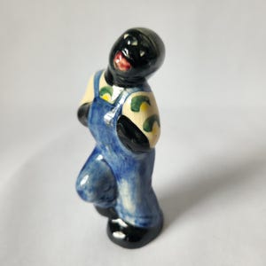 May include: A ceramic figurine of a person in blue overalls with black shoes and a black head. The figurine has a smiling expression with red lips and a cream-colored torso with green and yellow accents.