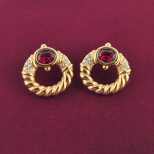 May include: A pair of gold-tone circular earrings featuring a twisted rope design. Each earring is centered with an oval-cut deep red gemstone and accented with small clear crystals along the upper curve of the circular frame.