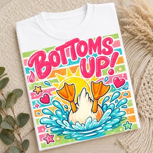 May include: White t-shirt with a colorful graphic featuring a duck diving into water, with the words "Bottoms Up!" in pink. The design includes a sun, hearts, and stars.